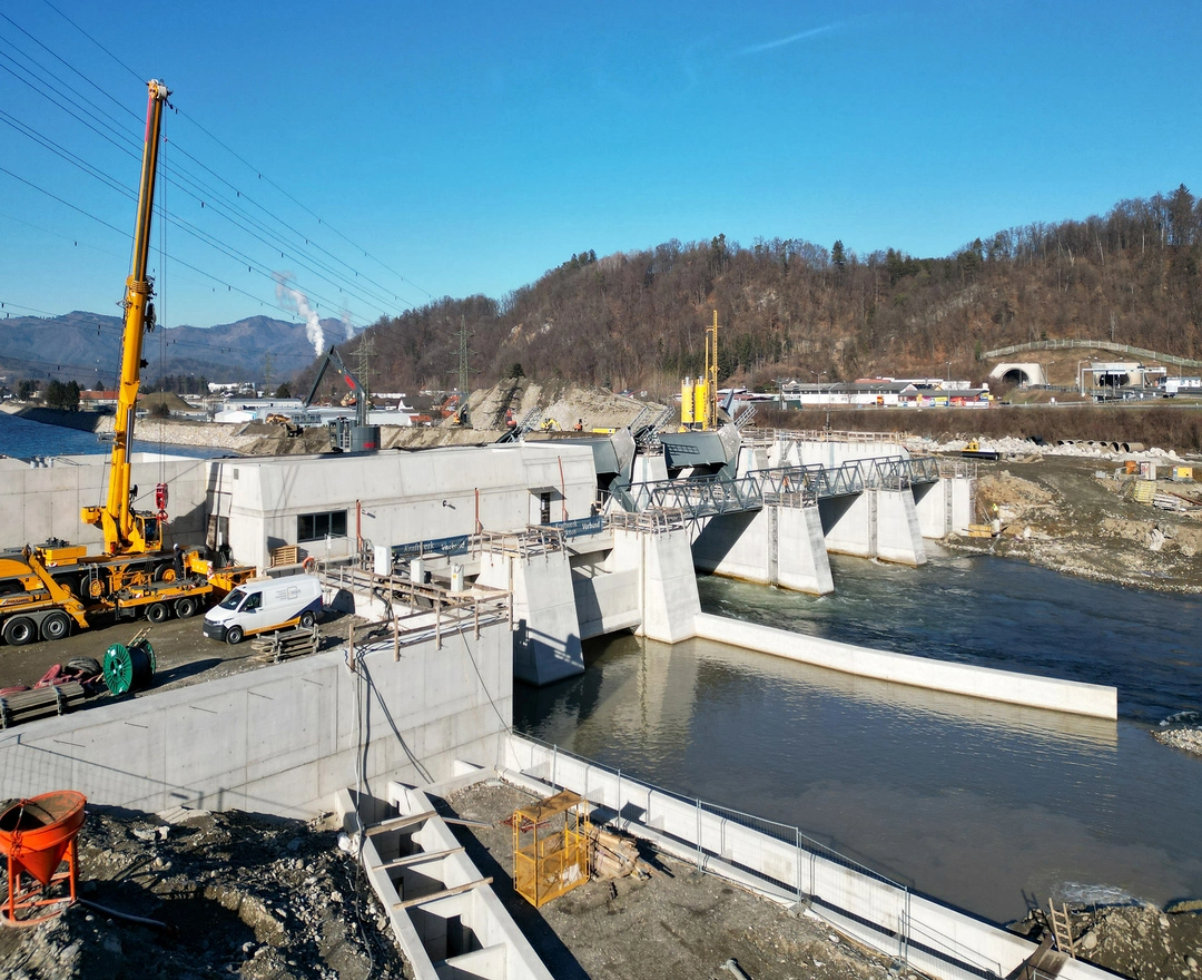 Blick auf die Baustelle des Wasserkraftwerks, mit Schwerpunkten auf der Konstruktion und den technischen Einrichtungen. Die Wehranlage mit mehreren geöffneten Schützen, welche die Wassermenge regulieren und leiten, ist deutlich zu erkennen. Im Vordergrund ist ein großer gelber Kran positioniert, der die Montage schwerer Bauteile unterstützt. Arbeitsfahrzeuge und Maschinen sind um die Baustelle verteilt, was auf eine rege Bautätigkeit hinweist. Im Hintergrund ist eine Hügellandschaft zu sehen, was die Integration des Kraftwerks in die natürliche Umgebung unterstreicht.