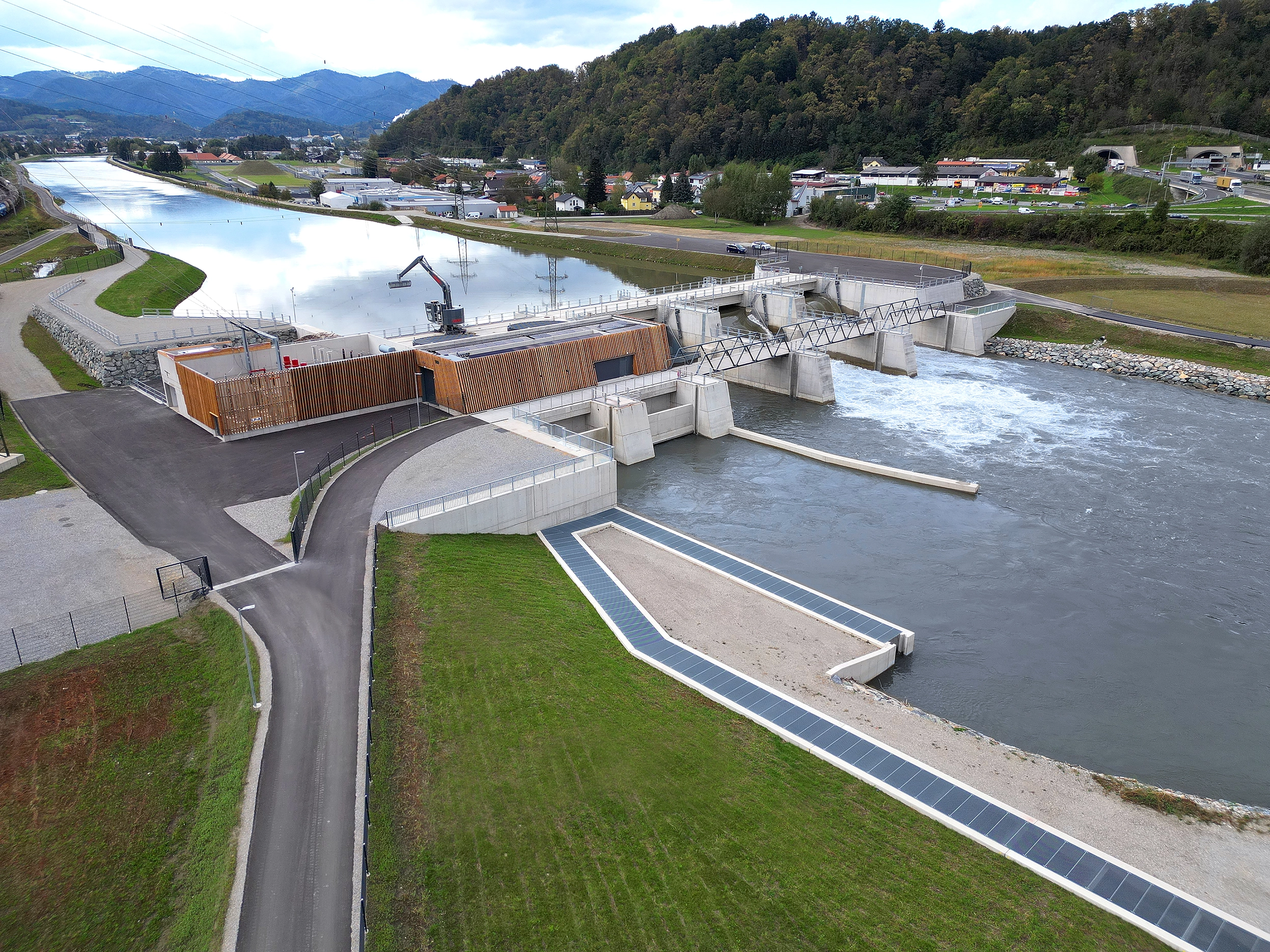 Wir blicken auf das neue Laufkraftwerk Gratkorn an der Mur. Die Fassade besteht aus einer Mischung aus Holz und Beton und wirkt sehr modern. Die Umgebung ist ländlich. Der Himmel leicht bewölkt.